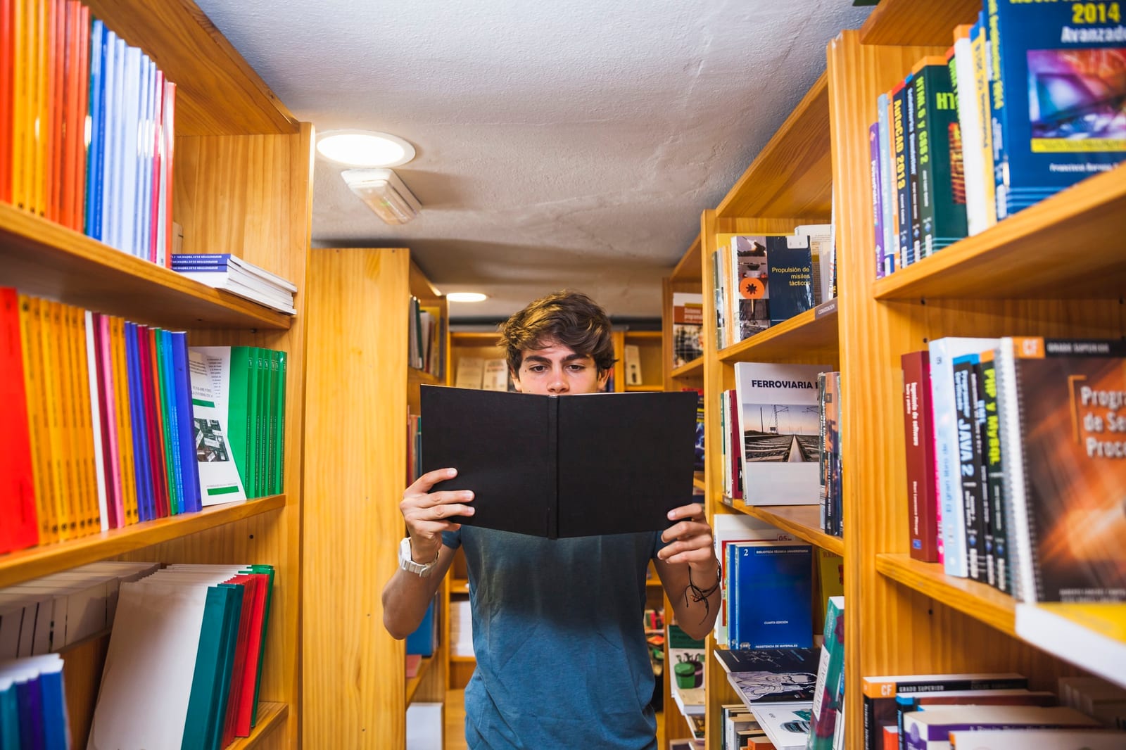 Jovem lendo atrás de um livro de capa preta em um corredor de biblioteca com estantes de livros técnicos