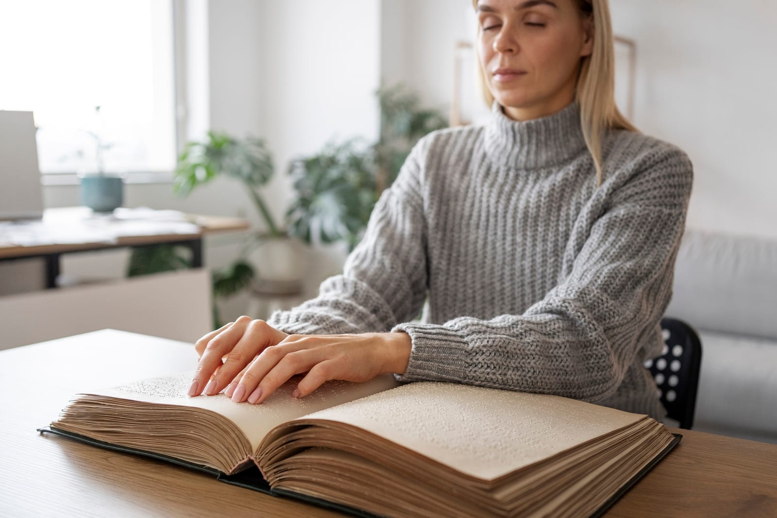 Mulher lendo um livro em braile com as pontas dos dedos, sentada a uma mesa com plantas ao fundo